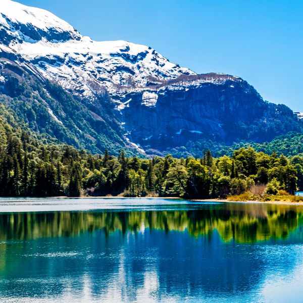 Snow-capped mountain reflecting in a clear blue lake surrounded by green forest.