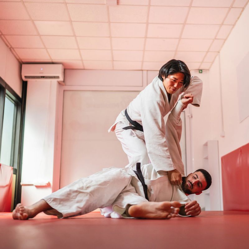 Two dedicated martial artists, a man and a woman, are practicing grappling techniques on a mat in a brightly lit dojo, showcasing their strength, skill, and discipline