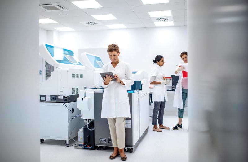 Mid adult Caucasian female lab professional wearing a white coat studies data on a tablet in a modern laboratory. Advanced equipment and diverse colleagues in the background highlight a collaborative scientific environment.