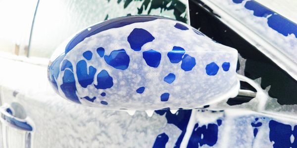 Blue car side mirror covered in white soap suds during a wash.