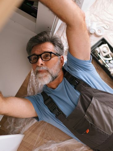 Man in glasses repairing something under a sink.