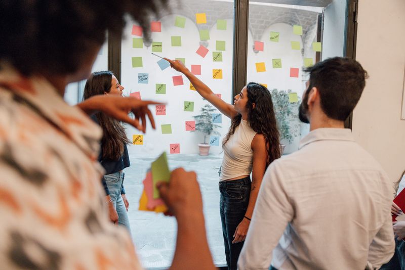 Three colleagues stand in front of a large glass wall covered with sticky notes, actively exchanging ideas. Their teamwork and engagement capture the essence of creative problem-solving in a modern business environment.