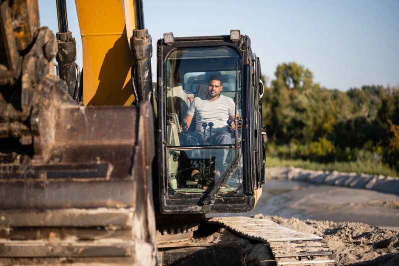 Construction worker operating heavy machinery under the bright sun at a bustling construction site, focused on driving the excavator