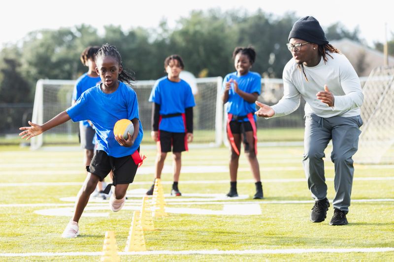 A coach with a multiracial group of players on a co-ed flag football team at practice. They are on the field doing drills. The focus is on the 8 year old girl, running with the football, zig-zagging through cones as her teammates watch and clap to encourage her.
