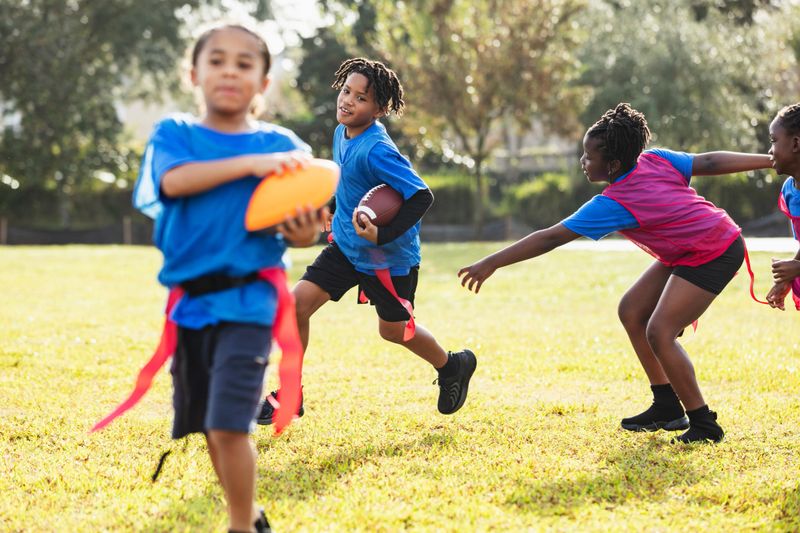 A multiracial group of children playing flag football in the park. The main focus is on the 10 year old Hispanic boy carrying a football.