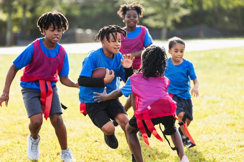 A multiracial group of five children playing co-ed flag football in the park. A 10 year old Hispanic boy is carrying the football, trying to avoid the opponents.