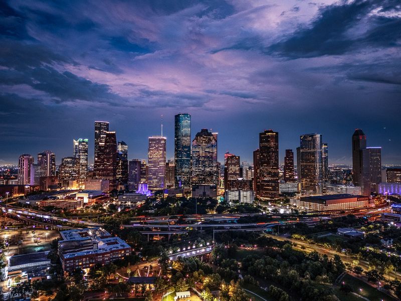 Drone aerial image of downtown Houston, Texas captured at twilight. The photograph highlights the illuminated skyscrapers, highways, and cityscape under a dramatic evening sky. A vibrant view of one of the largest cities in the United States and a major economic hub.