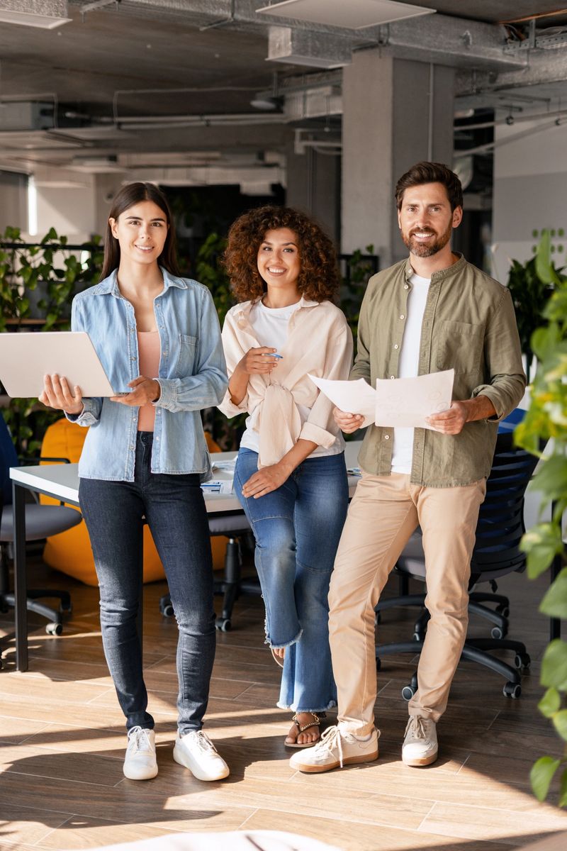 Three smiling colleagues are standing in their modern office, holding a laptop and documents, showcasing teamwork and collaboration in a startup environment. Teamwork concept