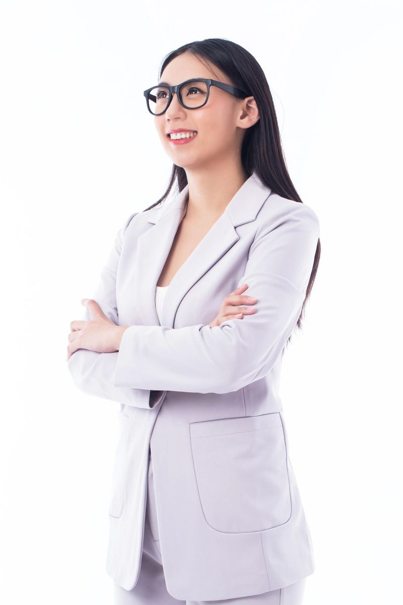 A confident young professional woman is standing with her arms crossed, smiling confidently. She is wearing a light gray blazer, a white top, and black glasses. Her long straight hair falls over her shoulders. The image is set against a clean white background, emphasizing her professional and approachable demeanor. She exudes positivity and determination, representing modern workplace confidence and empowerment.