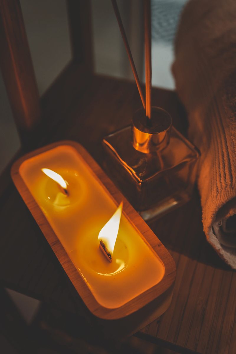 A handmade wooden candle with twin flames burning brightly on a bamboo shelf in a spa-style bathroom, accompanied by a rolled towel and glass diffuser, creating a serene and cozy atmosphere.