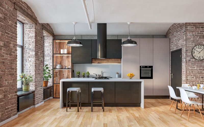 Modern industrial-style kitchen interior featuring exposed brick walls, sleek black and beige cabinets, and a central island with bar stools and with a built-in cooktop, on hardwood floor. Large windows allow natural light to brighten the space, complemented by indoor plants, open shelving, and stylish pendant lighting. A cozy dining area with white chairs and a minimalist table completes the contemporary design. 3D rendered image.