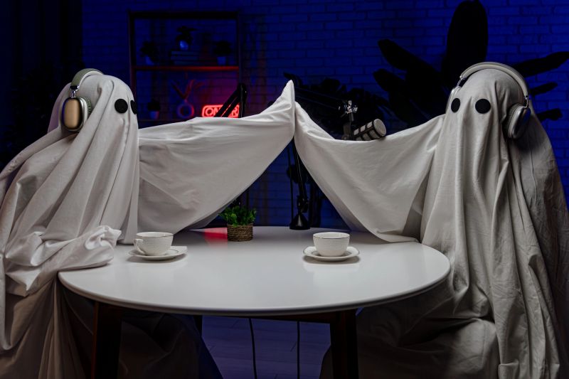 Two people dressed as ghosts, wearing white sheets and headphones, record a podcast at a studio table with microphones. Perfect for Halloween, comedy, and entertainment.