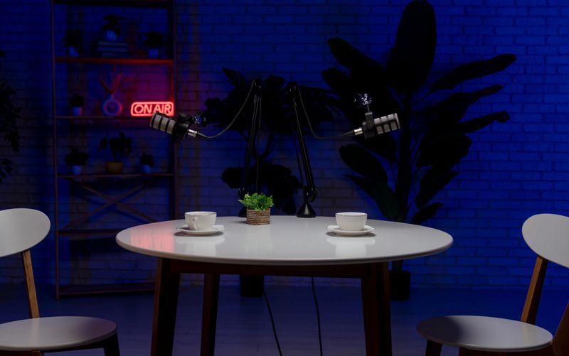 Modern podcast studio setup with round table, microphones, chairs, coffee cups, and neon ON AIR sign in the background. Perfect for media, broadcasting, and lifestyle themes.