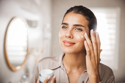 Woman applying skincare cream to her face in a bright room.