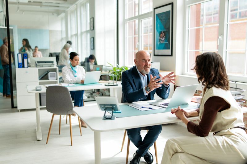 Experienced financial advisor providing guidance to a female customer while seated at his desk in a modern bank office.