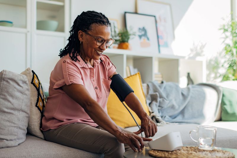 Black woman is seated comfortably on her living room sofa, carefully using a digital blood pressure monitor wrapped around her upper arm. She watches the display screen with focus, tracking her blood pressure and heart rate as part of her daily heart health routine