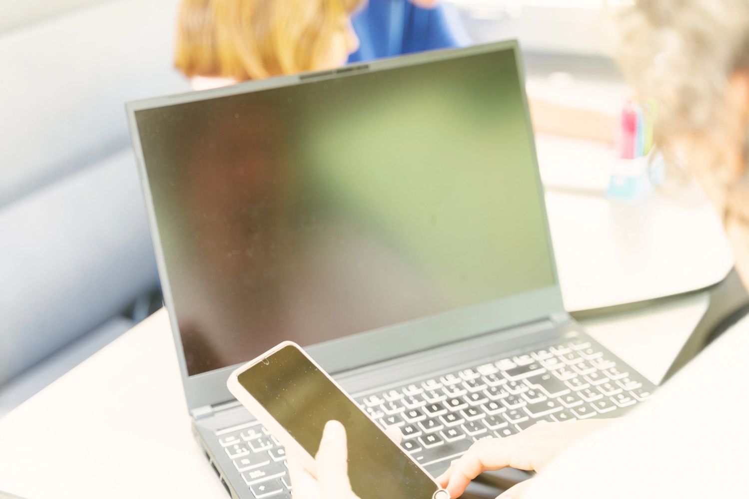 Person using a smartphone in front of a laptop with a blank screen.