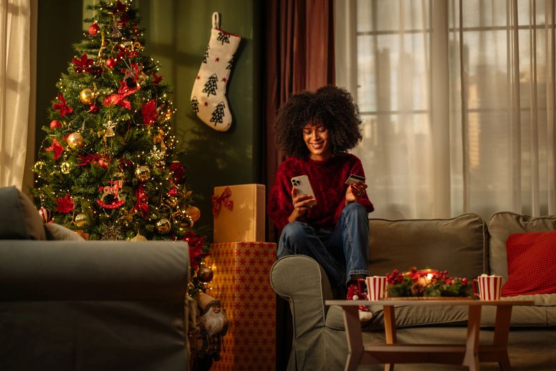 Young adult relaxing on a cozy sofa, using a smartphone and holding a credit card, with a beautifully decorated Christmas tree nearby