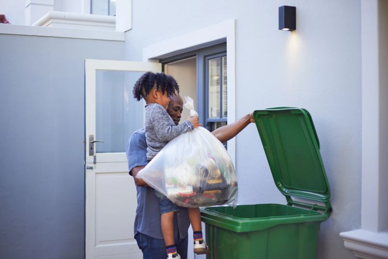 A heartwarming moment as a young boy is carried by his father or grandfather while helping take out the garbage or recycling. A simple, everyday scene showing care, responsibility, and togetherness across generations.