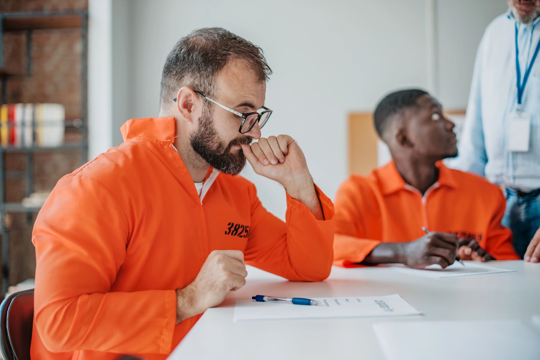 Men in orange prison uniforms sitting at a table, one looking thoughtful.