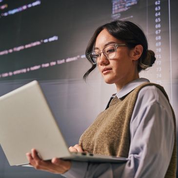 Woman working on a laptop with code displayed on a large screen behind her.