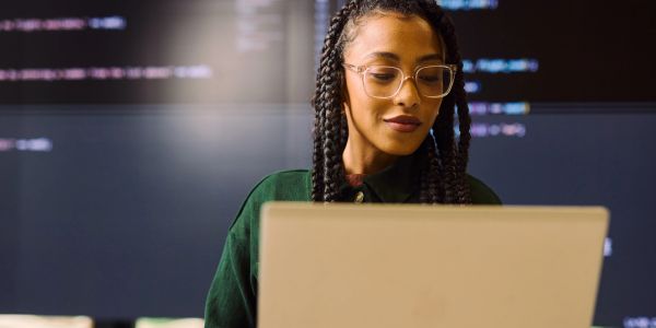 Woman working on laptop with code on screen behind her.