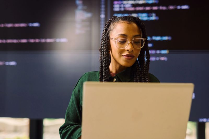 Young adult woman programmer analyzing code displayed on a large screen and laptop in a modern office