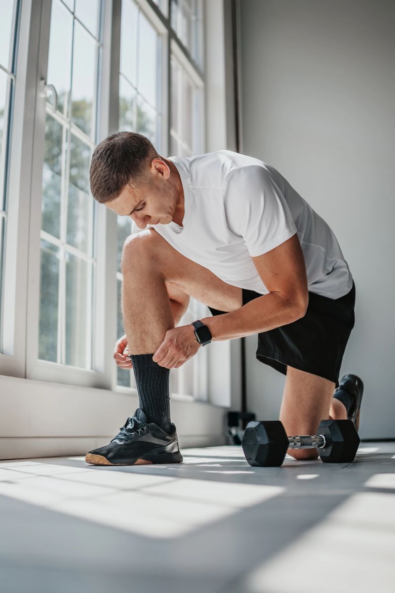 A young athletic man in a white sports shirt and black shorts kneels on the floor, tying his shoelaces next to a dumbbell.