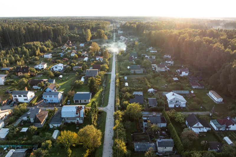 Aerial perspective of a residential area with houses along a straight road, surrounded by forest. Smoke rises from one property under warm sunlight.