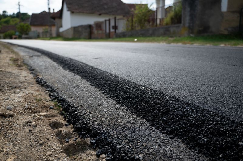 Close-up view of a newly paved asphalt road with a visible edge between the old and new surface. The scene is set in a quiet village with traditional houses lining the background. The image highlights road construction, infrastructure improvement, and rural development.