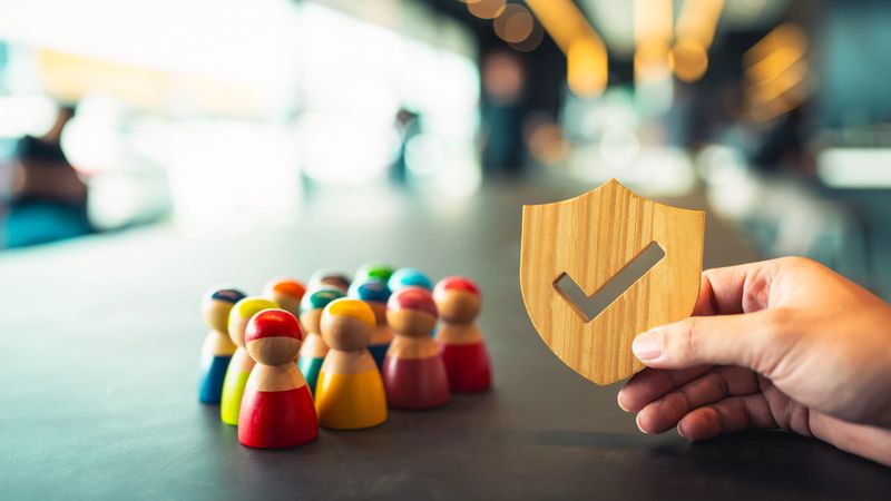 Wooden shield with checkmark protecting a diverse group of people figures. Concept of group insurance, employee protection, social safety, equality, diversity, HR benefits, and risk management.