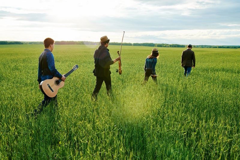 Group of young adult multiethnic musicians walking through green field carrying guitar and violin, moving away from camera, engaging in outdoor activity together in rural landscape