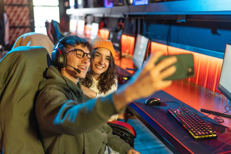 Two young adult gamers celebrating their victory in an esports tournament, joyfully taking a selfie with a smartphone in a vibrant internet cafe filled with colorful neon lights