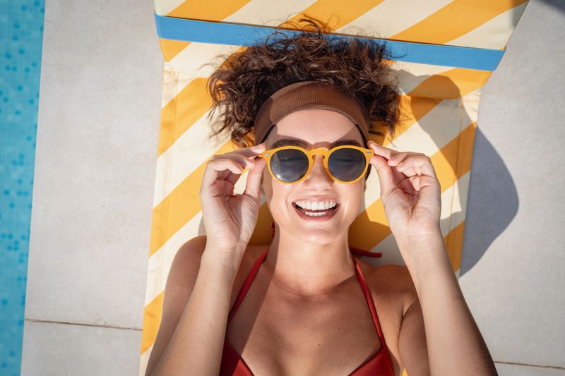 A woman smiles joyfully while reclining on a striped sun lounger next to a pool, enjoying her day.