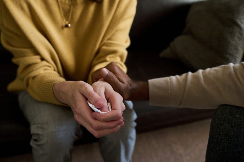 Caucasian young adult woman sitting with hands clasped holding tissue, Black middle aged man gently touching her arm offering comfort, both seated on sofa in indoor setting
