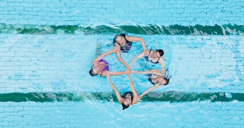 A overhead shot of a group of five senior women performing synchronized swimming inside a pool in an Edwardian bathhouse.