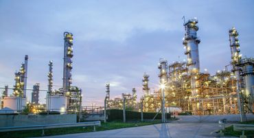 Illuminated industrial refinery towers at dusk under a cloudy sky.