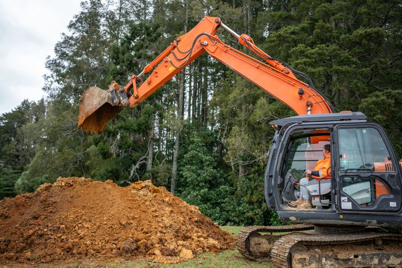 A worker operates a large orange excavator to lift and move soil