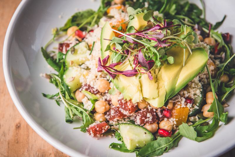 A vibrant and nutritious couscous salad featuring slices of avocado, mixed greens, pomegranate seeds, chickpeas, and cucumber. Perfectly balanced with fresh microgreens, creating a wholesome and delicious meal ideal for health-conscious individuals.