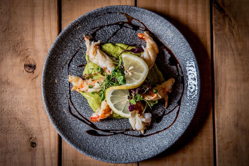 A gourmet dish featuring shrimp elegantly arranged on a bed of mashed avocado and drizzled with sauce, accented with fresh herbs and lemon slices, served on a textured plate. The rustic wooden table adds to the sophisticated presentation, making this image ideal for culinary, gourmet, or fine dining themes.