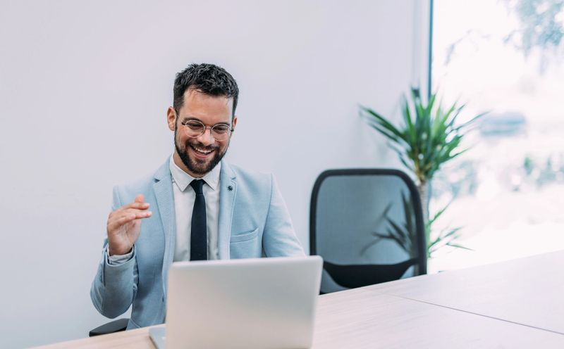 Handsome businessman having video call with his coworkers. Businessman having important online conversation with customer. Shot of a young man using a laptop for video conference. Smiling businessman in a video call using laptop in the office.