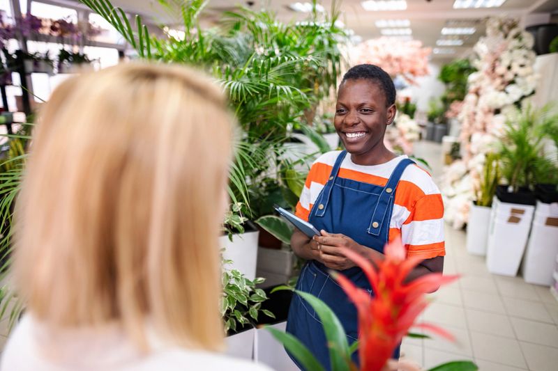 Florist holding tablet smiling at customer in flower shop, surrounded by plants