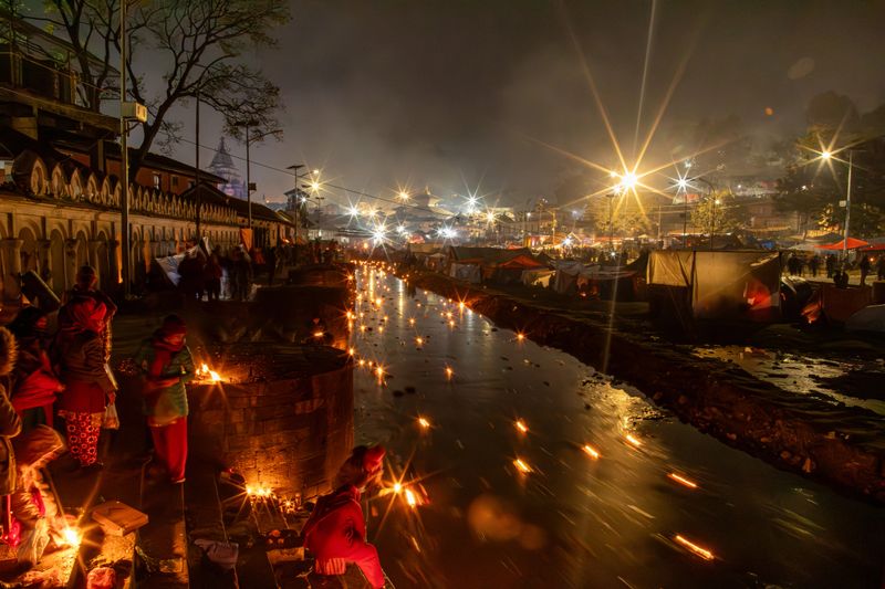 A breathtaking scene where devotees light and float oil lamps on the Bagmati River during a religious festival at night. The glowing lamps reflect off the water, creating a river of light. In the background, the famous Pashupatinath Temple can be seen under a starry sky. The event is serene and spiritual, with people paying homage and offering prayers for their loved ones.