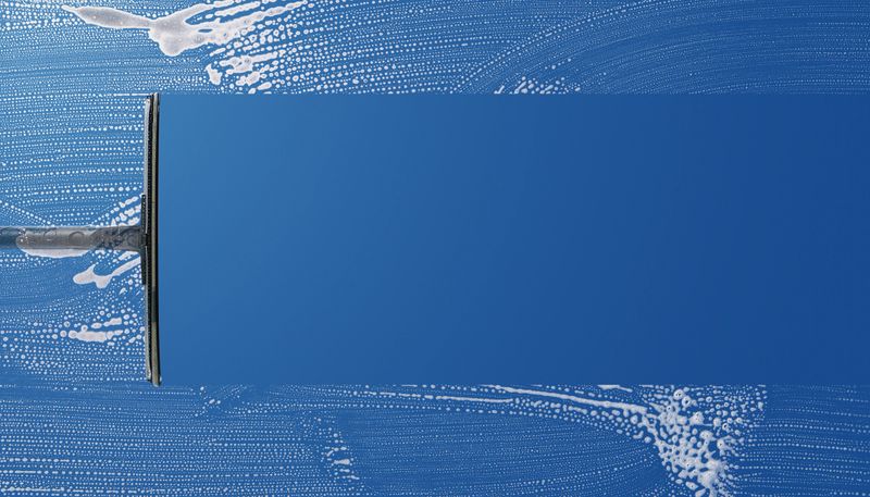 Close-up of squeegee wiping away soap foam from a glass window, leaving a clear surface with blue sky behind
