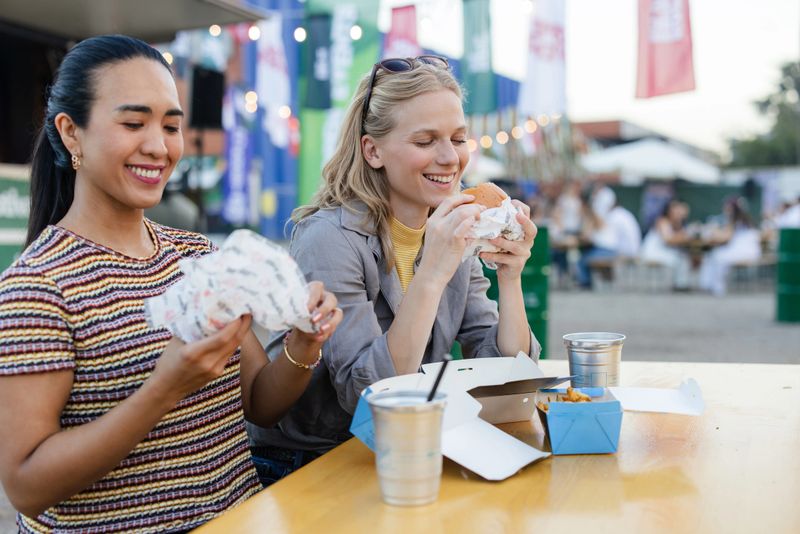 Two women share a joyful moment at an outdoor festival while enjoying delicious food under colorful decorations, creating a cheerful and lively atmosphere perfect for bonding and celebration.