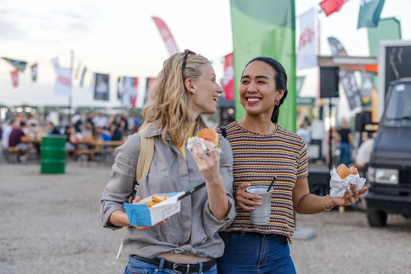 Two cheerful women share food and smiles at a lively outdoor festival setting, surrounded by vendor flags and food trucks, enjoying a casual and enjoyable day together outdoors.