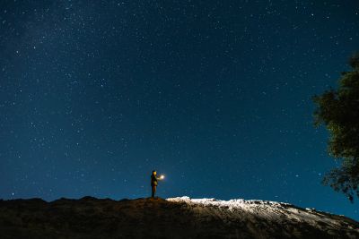 Person standing on a hill under a starry night sky holding a light.