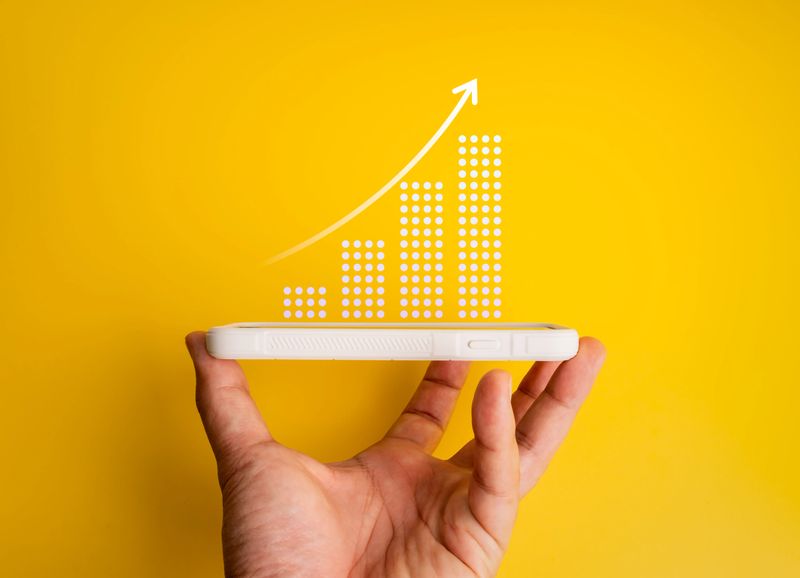 Hand holding smartphone with rising bar graph and arrow on yellow background, representing business growth, online investment, fintech, mobile marketing, digital success, and creative data strategy.