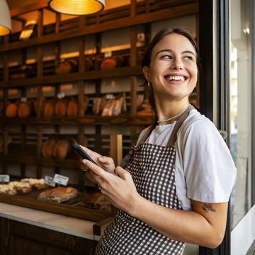 Smiling bakery worker holding a phone, standing by the window.
