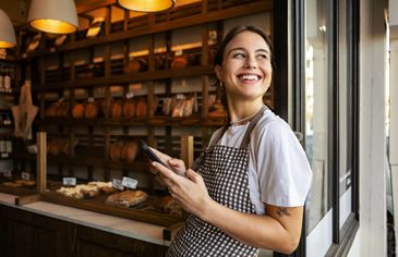 Smiling woman in apron holding phone in bakery.